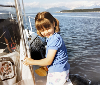 Young girl stands on a boat, water and mountains can be seen in the background.